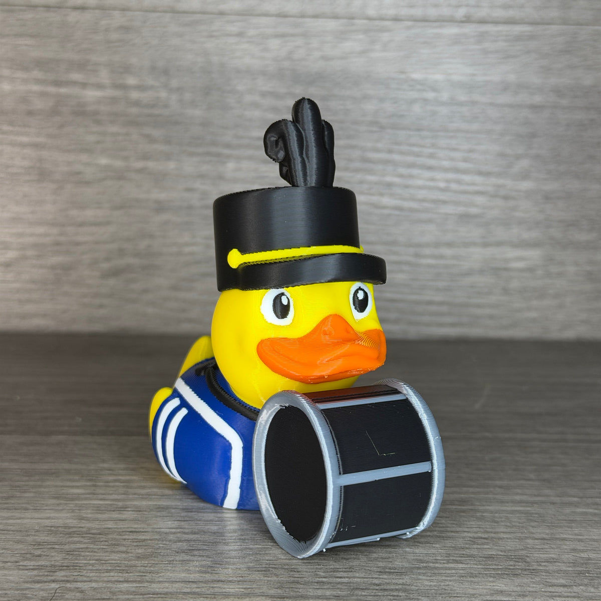 Yellow 3D printed duck dressed as a marching band drummer in a blue uniform with white trim, tall black plumed band hat, holding a black and silver bass drum, three-quarter front view