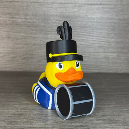 Yellow 3D printed duck dressed as a marching band drummer in a blue uniform with white trim, tall black plumed band hat, holding a black and silver bass drum, three-quarter front view