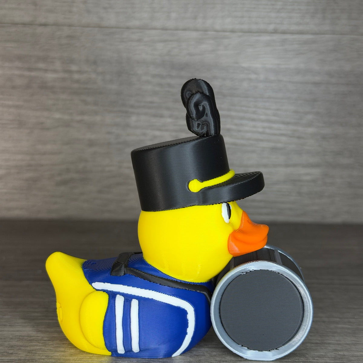 Side profile of the drummer duck showing the tall black band hat, curled plume, blue jacket stripes and bass drum