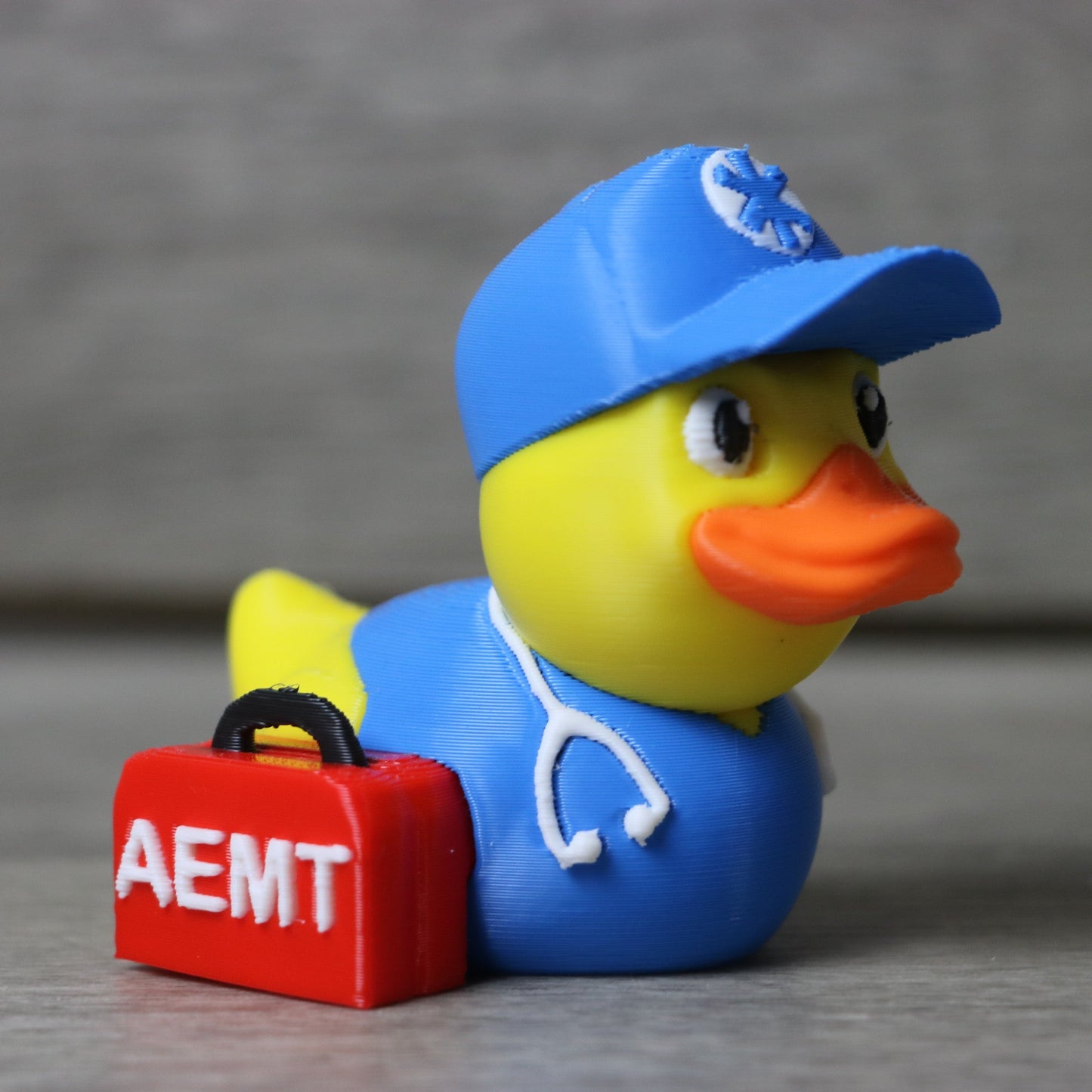 Rubber duck wearing a blue cap and holding a red suitcase with 'AEMT' on it, on a gray background.
